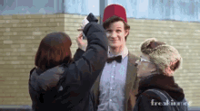 a woman is taking a picture of a man wearing a fez hat and a bow tie