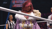 a woman in a purple dress stands in a wrestling ring with a aew logo on the screen behind her