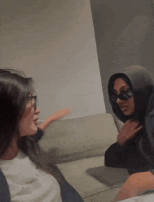 two women sitting on a couch with one wearing a hoodie and the other wearing sunglasses