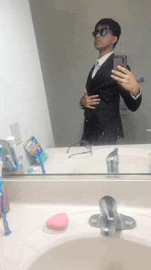 a man in a suit takes a selfie in front of a mirror with a tube of toothpaste on the counter