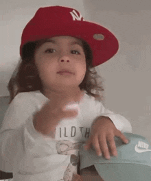 a little girl wearing a red hat and a white shirt is sitting on a bed .
