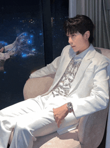a man in a white suit sits in a chair looking out a window