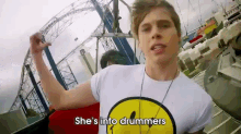 a man wearing a smiley face shirt says she 's into drummers in front of a roller coaster