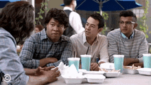 a group of men are sitting at a table with containers of food and drinks with a c on the bottom right