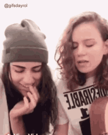 two girls are standing next to each other and one is wearing a beanie and the other is wearing a t-shirt .