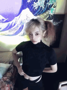 a girl in a black crop top stands in front of a painting of waves