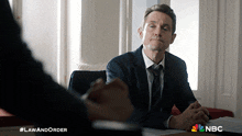 a man in a suit and tie is sitting at a table with a nbc logo in the background