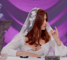 a woman in a white dress with a veil on her head is sitting at a table