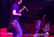 a man is singing into a microphone while a woman sits on a red couch behind him