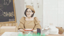 a woman wearing glasses sits at a table in front of a blackboard that says smile
