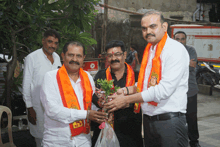 a man in a white shirt is giving flowers to another man wearing an orange sash