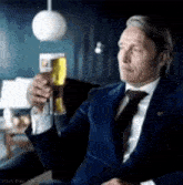 a man in a suit and tie is holding a glass of beer while sitting on a couch .