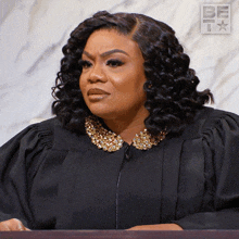 a woman wearing a black gown with a gold collar is sitting at a podium with a be i logo behind her
