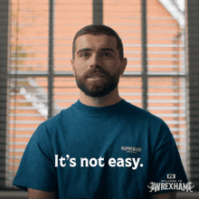 a man with a beard wears a blue shirt that says it 's not easy