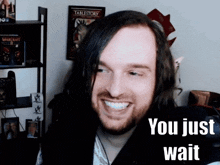 a man with long hair and a beard smiles with the words you just wait below him