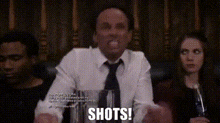 a man in a tie is sitting at a table with a glass of wine and the words `` shots '' written on the screen .