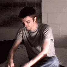 a man in a grey shirt is sitting on a bed with his eyes closed