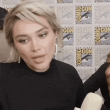 a woman talks into a microphone while standing in front of a comic con wall