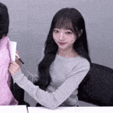 a girl with long black hair is sitting at a table holding a marker