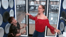 a woman in a red off the shoulder top is dancing with another woman in front of a microphone