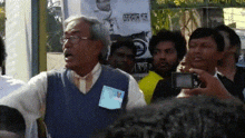 a man wearing glasses stands in front of a crowd with a sign in the background that says ' bangladesh ' on it