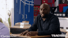 a man in a black shirt is sitting at a desk in a classroom with the word teachers on the bottom right