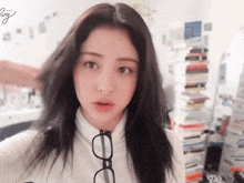 a woman wearing glasses and a white sweater looks at the camera with a stack of books behind her