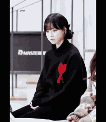 a girl wearing a black sweater with a red heart on the front sits in front of a sign that says master
