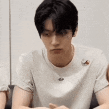 a young man wearing a white t-shirt is sitting at a table with his hands folded .