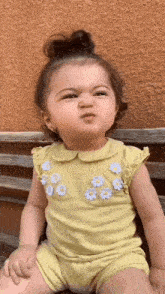 a little girl in a yellow dress is sitting on a bench making a funny face .