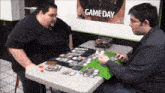 two men sit at a table playing a game with a game day poster behind them