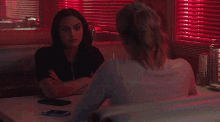 two women are sitting at a table with their arms crossed in front of a window with red blinds .