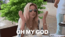 a woman wearing sunglasses and a necklace is sitting at a table with her hands in the air .