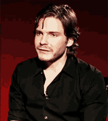 a man wearing a black shirt is sitting in front of a red wall