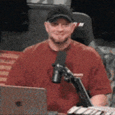 a man in a red shirt is sitting in front of a microphone in front of an apple laptop