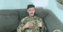 a soldier in a camouflage uniform is sitting on a couch holding a cell phone .