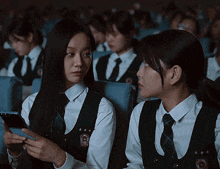 two girls in school uniforms are sitting in a theater looking at a cell phone