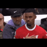 a man in a red shirt is standing next to a man in a black hat and holding his hand to his chest .