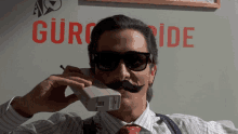 a man with a fake mustache is talking on a phone in front of a sign that says gürc ide