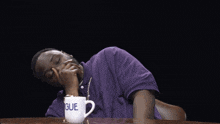 a man in a purple shirt is sitting at a table with his arms outstretched in front of a mug that says gue