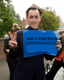 a man in a suit holds up a sign that says shut the fuck up