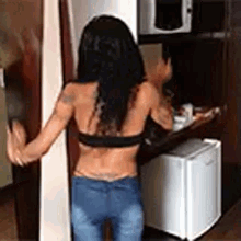 a woman in a bra and jeans is standing in front of a refrigerator in a kitchen .