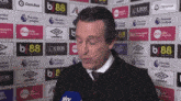 a man is talking into a microphone in front of a wall covered in advertisements for premier league and umbro