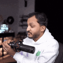 a man in a white hoodie is smoking a cigarette while holding a camera in his hands .